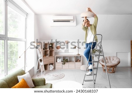 Young man standing on ladder and changing light bulb in lamp at home