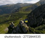 Young man standing on sharp rocky ridge holding blue and yellow Ukrainian flag. Drone shot of hiker on Shpytsi peak in Chornohora range. Patriotic concept and mountain adventure in Ukraine.
