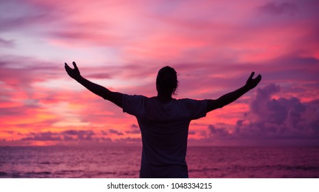 young man is standing on the shore of the ocean against the background of an incredible purple sunset, flinging his hands to the side - Powered by Shutterstock