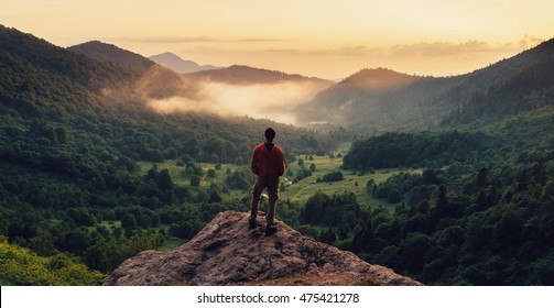 Young man standing on top of cliff in summer mountains at sunset and enjoying view of nature