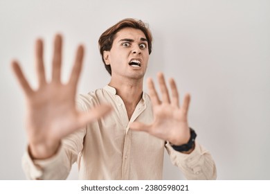 Young man standing over isolated background afraid and terrified with fear expression stop gesture with hands, shouting in shock. panic concept. 
