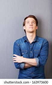 Young man standing thinking with his arms folded and head tilted back looking up into the air on a grey background