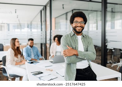 Young man stands with arms crossed in modern office space, showcasing relaxed yet professional work environment, behind him diverse team of colleagues engages in a productive and collaborative meeting - Powered by Shutterstock