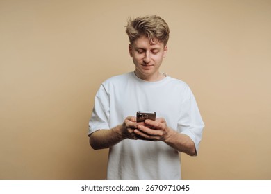 A young man stands casually while interacting with his smartphone. He appears attentive, showcasing a relaxed posture against a simple, neutral backdrop.