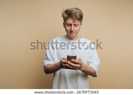 A young man stands casually while interacting with his smartphone. He appears attentive, showcasing a relaxed posture against a simple, neutral backdrop.
