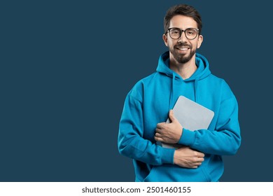 A young man stands confidently with a smile, wearing a blue hoodie and holding a laptop closely to his chest. The background is a dark color, copy space - Powered by Shutterstock