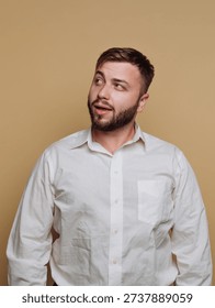A young man stands with a curious expression, wearing a crisp white shirt. His beard adds to his charm as he looks upward against a soft beige backdrop, evoking a sense of contemplation.