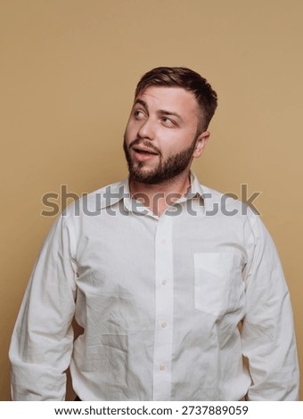 A young man stands with a curious expression, wearing a crisp white shirt. His beard adds to his charm as he looks upward against a soft beige backdrop, evoking a sense of contemplation.