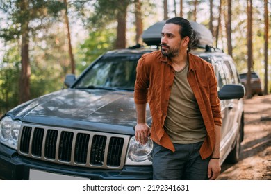A young man stands next to a car with an upper tourist trunk in a pine forest on the shore of a lake at sunset. A handsome guy is a tourist and an SUV among the trees on a forest road in the sunlight. - Powered by Shutterstock