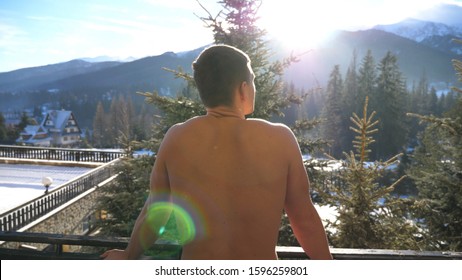 Young Man Stands On Balcony And Breathes Fresh Air During Sunny Day. Athletic Guy Enjoys Beautiful Mountain And Forest Landscape In Morning. Concept Of Winter Vacation Or Holiday. Dolly Shot Slow Mo