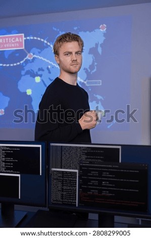 A young man stands in a technology-centric workspace, surrounded by monitors displaying technical data and global analytics. He is part of an operations center team analyzing cybersecurity threats. The environment reflects a focus on real-time threat management and advanced data analytics, emphasizing the importance of modern technology in safeguarding information systems.