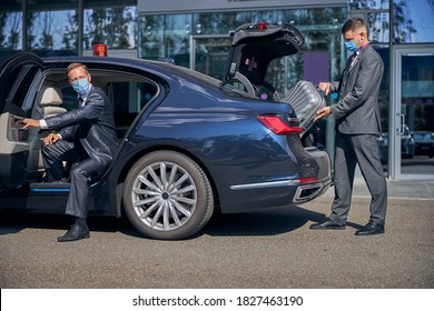 Young Man In Sterile Mask Is Packing Boss Luggage Into Trunk While He Is Getting Into Car