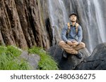 young man stops to sit and meditate during a hiking session in front of a large waterfall in the middle of nature, peace of mind, meditation, serenity