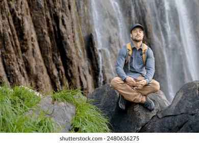 young man stops to sit and meditate during a hiking session in front of a large waterfall in the middle of nature, peace of mind, meditation, serenity - Powered by Shutterstock