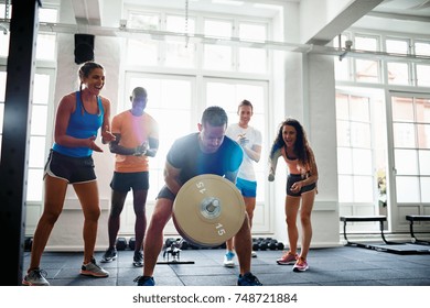 Young man straining to lift a barbell in a gym with a diverse group of friends cheering him on in the background