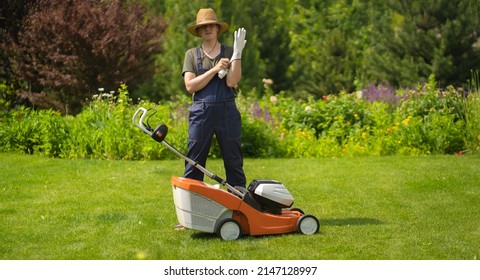 A young man in a straw hat is putting on gloves to mowe a lawn with a lawn mower in his beautiful green floral summe garden. A professional gardener with a lawnmower cares for the grass.