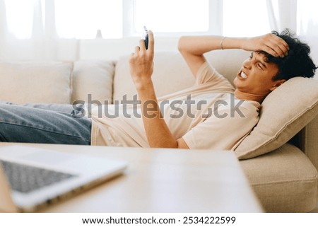young man stressed while using smartphone, sitting on a couch, casual outfit, indoor setting with natural light, frustration and technology reliance theme