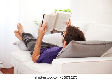 Young Man Stretching Comfortably On Couch And Reading A Book. Man Relaxing At Home