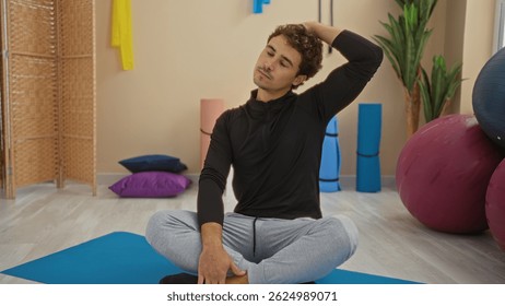 Young man stretching neck in an indoor gym setting, showcasing relaxation and fitness in a sports environment, surrounded by exercise mats and equipment.