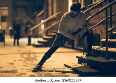 Young man stretching while out jogging and exercising at night during winter and snow in the city - Powered by Shutterstock