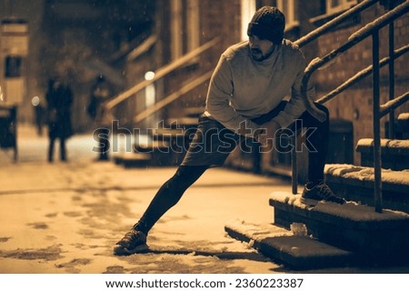 Young man stretching while out jogging and exercising at night during winter and snow in the city