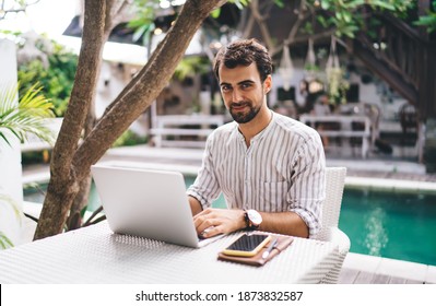 Young Man In Striped Shirt And Wristwatch Looking At Camera While Working On Laptop In Bright Outside Cafe Near Swimming Pool