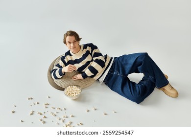 A young man in a striped sweater lounges comfortably, savoring popcorn in a studio setting. - Powered by Shutterstock