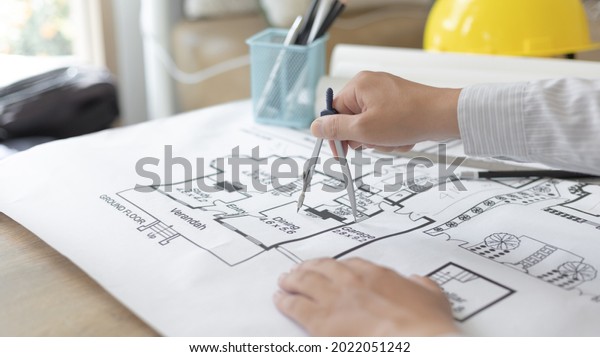 Young man  structural design or engineer is\
using a measuring circle around a floor plan or blueprint,\
Architect or engineer is designing a building using compasses to\
draw the physical\
structure.