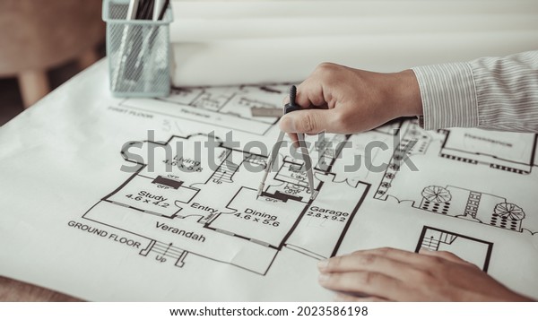 Young man  structural design or engineer is\
using a measuring circle around a floor plan or blueprint,\
Architect or engineer is designing a building using compasses to\
draw the physical\
structure.