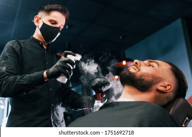 Young Man With Stylish Hairstyle Sitting And Getting His Beard Shaved By Guy In Black Protective Mask In Barber Shop.