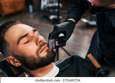 Young Man With Stylish Hairstyle Sitting And Getting His Beard Shaved By Guy In Black Protective Mask In Barber Shop.