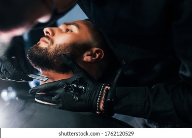 Young Man With Stylish Hairstyle Sitting And Getting His Beard Shaved By Guy In Black Protective Mask In Barber Shop.