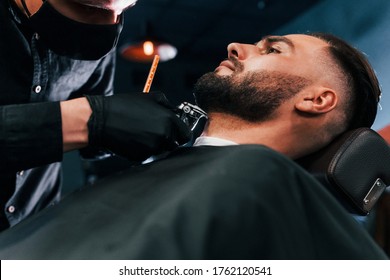 Young Man With Stylish Hairstyle Sitting And Getting His Beard Shaved By Guy In Black Protective Mask In Barber Shop.