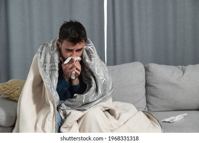 Young man suffering from a common cold and flu or allergy sit at home wrapped in blanket and wipe his nose with tissues while he have a strong headache pain, healthcare concept - Shutterstock ID 1968331318
