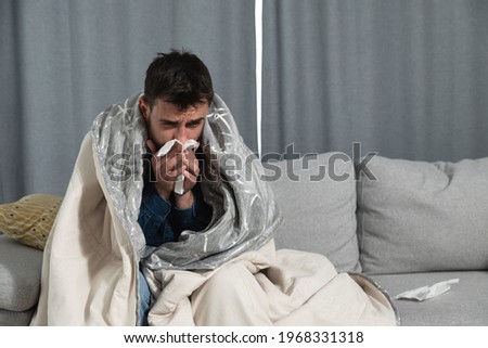 Young man suffering from a common cold and flu or allergy sit at home wrapped in blanket and wipe his nose with tissues while he have a strong headache pain, healthcare concept
