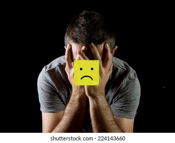 Young Man Suffering Depression And Stress Sitting Alone In Pain And Grief Feeling Desperate With Yellow Note Sad Face Stuck On His Hands Isolated On Black Background