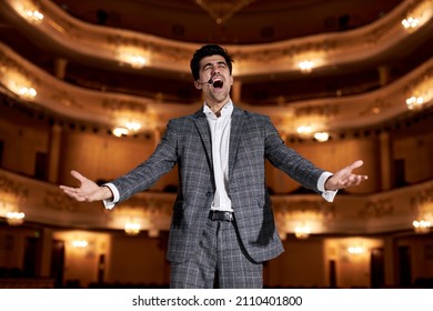 Young Man In Suit Gesticulating And Singing To Audience In Theater. Singer Hold Performance To Audience People. Presentation Stage. Unidentifiable Audience And Caucasian Singer Raising Hands