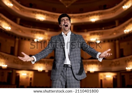 young man in suit gesticulating and singing to audience in theater. Singer hold performance to Audience People. Presentation Stage. Unidentifiable Audience and Caucasian Singer Raising Hands