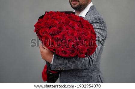 Young man in a suit holds a large bouquet of red roses on his shoulder, clenching his fist like a macho, because he wants to propose to his girlfriend.