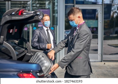 Young Man In Suit And Mask Packing Businessman Luggage Into Car Trunk After His Arrival To Airport