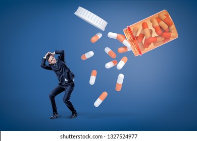 A Young Man In A Suit Trying To Protect Himself From An Avalanche Of Enormous Two-colored Pills Coming Out Of A Pill Jar. Pharmaceutical Companies Are Expanding Distribution Markets. Drug Tolerance