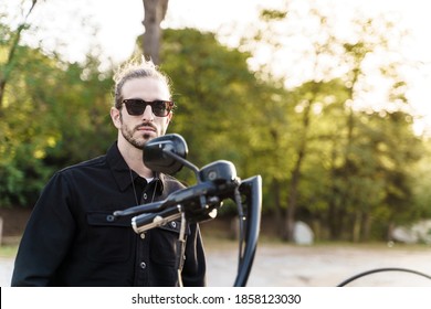 Young Man With Sunglasses And Rude Expression In Front Of A Motorcycle In A Parking Lot Surrounded By Trees