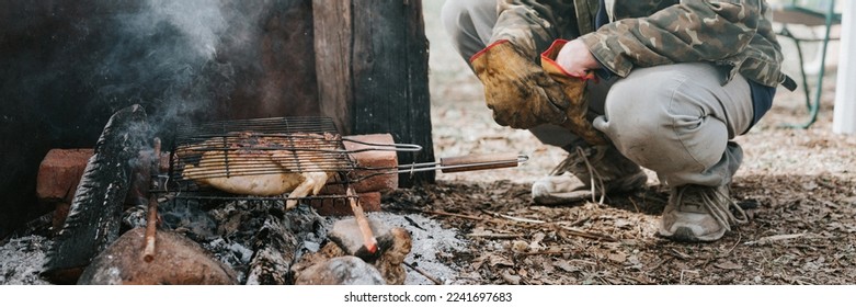 young man survivalist cooks roasts chicken meat food are fried on grill on smoldering coals or ember from campfire on ground. barbecue in camping conditions. countryside and wild rustic life. banner - Powered by Shutterstock