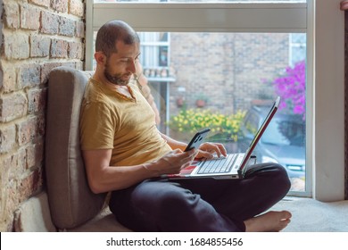 Young Man In Sweat Pants And T-shirt Working At Home With His Laptop On His Leg While Look His Smartphone