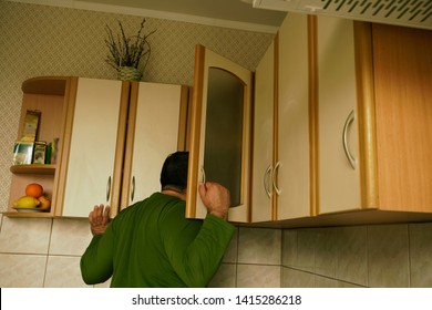 Young man in sweater peeks into kitchen cupboard at home, viewed from the back side. alone hungry man search food. 