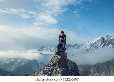 A young man in a sweater with phone in hand on the top of a snowy mountain far from civilization on a background of blue sky. The concept of activity and the availability of mobile connection