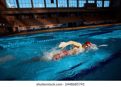 Young man, swimmer in red cap and goggles in motion, training freestyle stroke, swimming in pool indoors. Concept of professional sport, health, endurance, strength, active lifestyle - Powered by Shutterstock