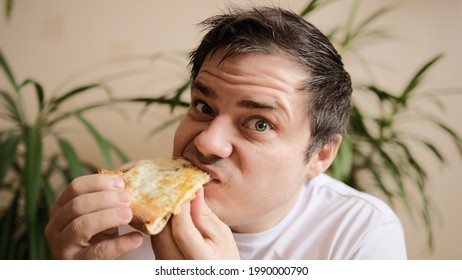 The Young Man Takes A Slice Of Pizza, Takes A Bite With Passion And Chews It. The Guy Looks At A Slice Of Pizza Adoringly. Delicious Cheesy Pizza. Green Plants On The Background.