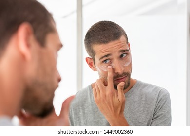 Young man taking care of his undereye wrinkles putting anti aging eye moisturizer. Handsome guy applying moisturizer and looking at himself while standing in front of the mirror in the bathroom.  - Powered by Shutterstock