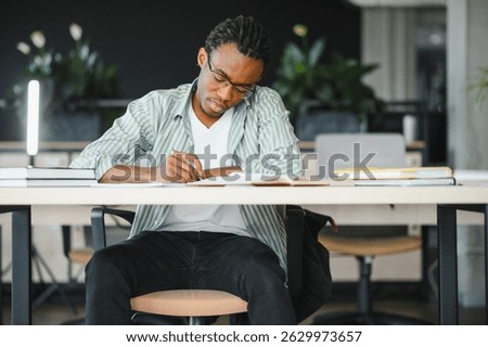Young man taking notes from textbook, preparing exam at university library, focused student learning and studying